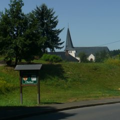 Blick auf die mit Bäumen umfasste Pfarrkirche am Ortseingang mit Begrüßungsschild
